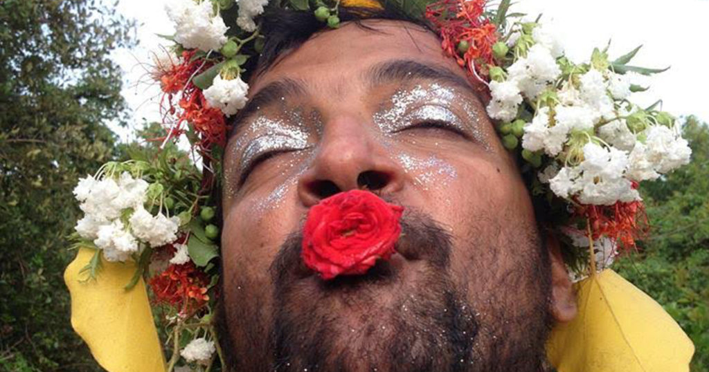 Anuj's face as he lays in a field of flowers, with silver eyeshadow and a red rose on his lips.