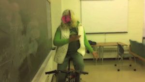 Anuj in his MIss Piggy mask riding a stationary bike in a classroom.