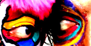 A close up of two faces, each covered in colorful make-up, looking at each other.