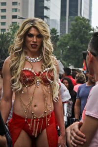 A gender performer in a long, curly blond wig wearing a red bikini with several layers of beads draped over them.