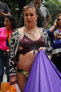 A gender artist wearing a bikini top designed to resemble a purple butterfly, a black and white jacket, and holding a purple cloth.