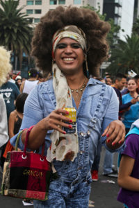 An individual in an all-denim disco outfit smiles into the camera while holding a drinkl, showcasing their pink nails which match their pink purse.