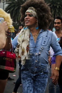 A candid shot of an individual wearing large hair tied back in a long headbnad, a denim button-down, and denim overalls.