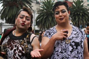 Two indiviuals in pedestrian clothing, one of whom blows a kiss to the camera. One wears a cheetah-print top and the other wears a black-and-white floral top.