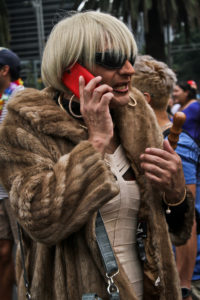 An individual wearing a heavy fur coat, a light colored dress, a short blond bob and with a red cell phone to their ear.