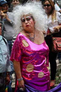 A gender artist wearing bright and exaggerated make-up, a large white wig, and a low-cut, shiny pink dress with three large yellow polka dots.