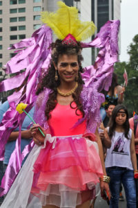 An individual wearing a bright pink dress with large, flowing purple wings on and a yellow crown-like headpiece smiles at the camera.