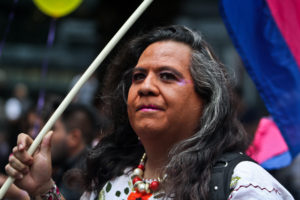 An individual smiling slightly while looking off camera, wearing long black hair and minimal make-up, while holding a rainbow flag.
