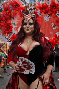 A gender artist dressed in a red one-piece with a flowing robe, red feathers, and several different playing cards attached.