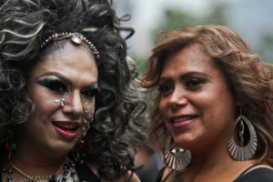 Two individuals looking at the camera while facing each other. One wears big curly black hair and heavy make-up, including many facial rhinestones, while the other wears feathered brown hair and large earrings with minimal make-up.