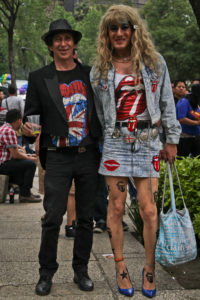 Two individuals each wearing Rolling Stones t-shirts. One wears short hair and black clothes, while the other wears a denim outfit with a short skirt and long blond wig.