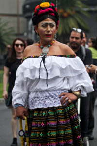 An individual wearing black hair in an updo fastened with colorful flowers, a shoulderless white top, and a black, green, and pink patterned skirt.