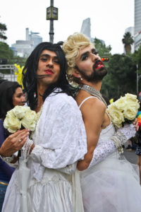 Two gender performers in white wedding gowns, standing back to back, looking near the camera while holding pale yellow roses. One wears a think brown beard and the other is cleanshaven.