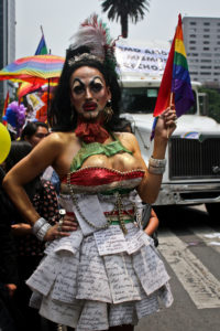 A drag performer dons clownish make-up and a dress that appears to be made out of the pages of books.
